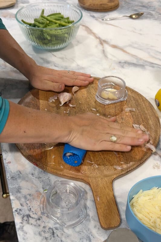 Crystel peeling garlic cloves with a silicone garlic roller on a wooden board, with trimmed green beans nearby.