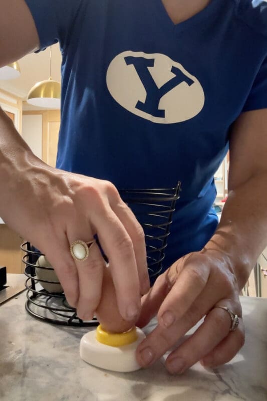 Close up of Crystel pressing a fresh egg down onto an egg piercer on the counter, poking a small hole in the wide end to test whether it helps hard boiled eggs peel more easily.