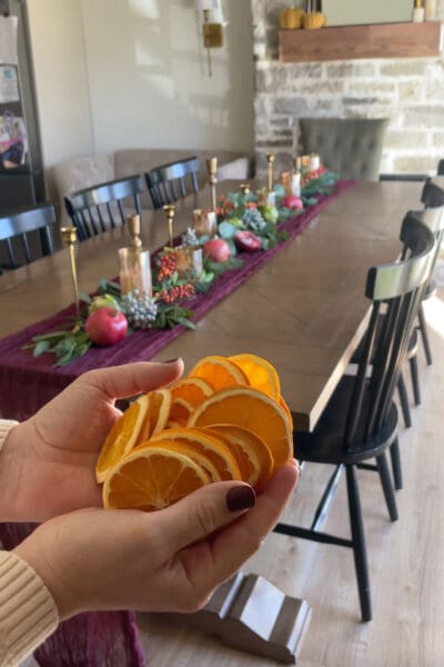 Hands holding a stack of dried orange slices above a burgundy centerpiece of pomegranates, berries, and eucalyptus, the detail layer that makes a seasonal tablescape feel like fall