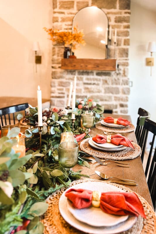 Long dining table set with a terracotta velvet runner, eucalyptus and pine garland, brass bowls of faux mums, ivory taper candles, and terracotta napkins in gold rings, showing seasonal tablescape ideas for a holiday dinner