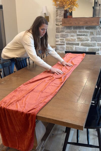 Crystel smoothing a terracotta velvet runner down the center of a wooden dining table, the first layer in building any seasonal tablescape