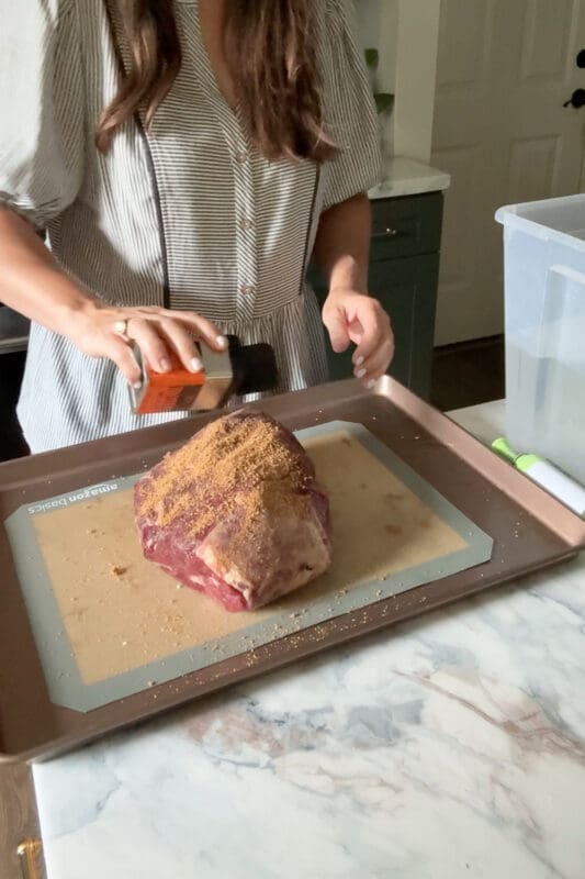 Crystel sprinkling Traeger rub over a raw chuck roast on a baking sheet to prep it for the sous vide method
