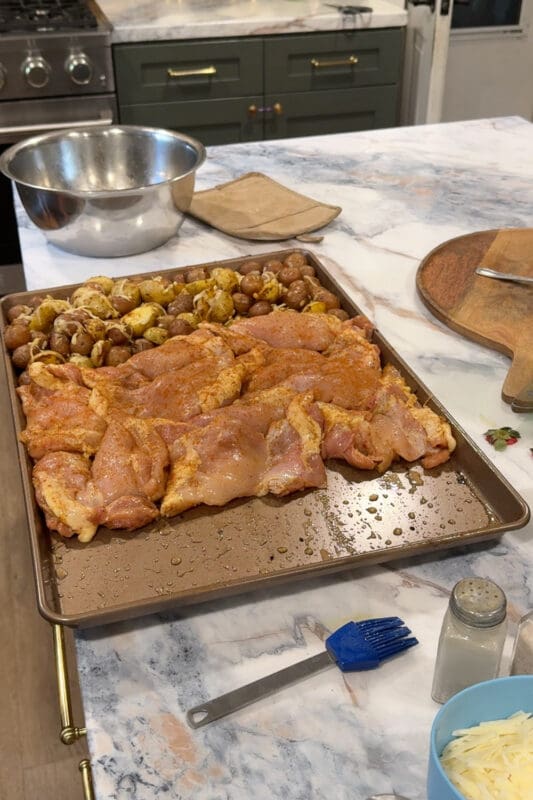 Seasoned raw chicken thighs arranged on a sheet pan beside parmesan baby potatoes, ready to roast for sheet pan chicken thighs.