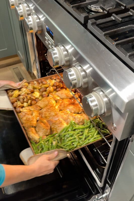 Crystel pulling a finished sheet pan of chicken thighs, roasted potatoes, and garlicky green beans from the oven.