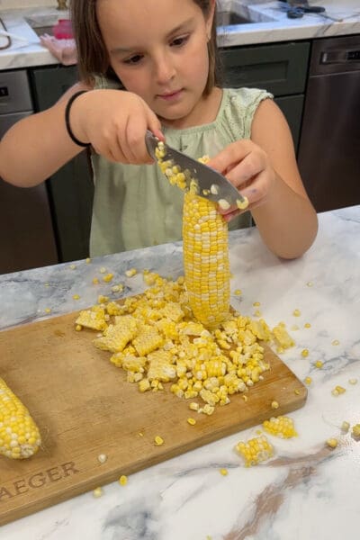 A young helper cutting fresh corn kernels off the cob onto a wooden cutting board for shrimp and corn succotash