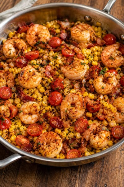 Close up of shrimp and corn succotash topped with torn herbs and bacon, served in a skillet on a wood table