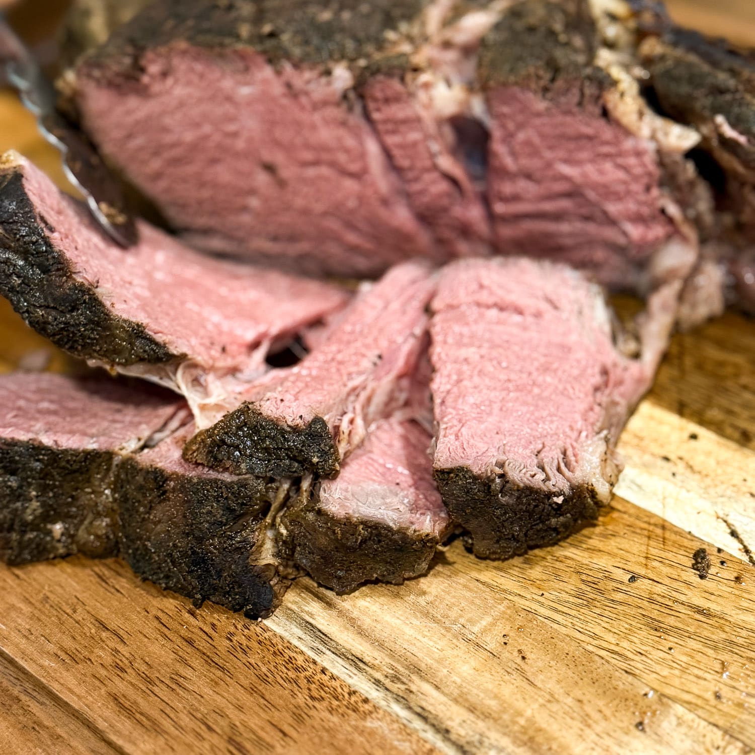 Tongs lifting a slice from a sous vide chuck roast on a wood cutting board, showing the pink interior and dark seasoned crust