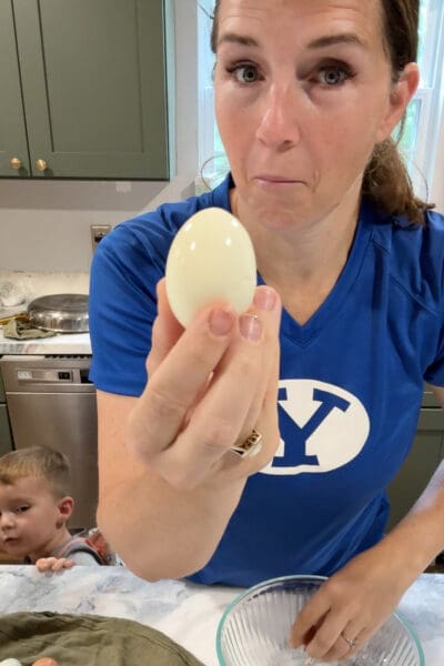 Crystel holding up a smooth, cleanly peeled hard boiled fresh egg with no torn spots, the successful result after the ice bath, while her young son watches from the counter.