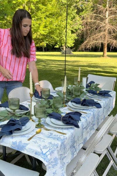 Crystel setting an outdoor table on the lawn with a blue toile tablecloth, navy napkins in gold rings, green ribbed goblets, and a eucalyptus garland, easy summer seasonal tablescape ideas