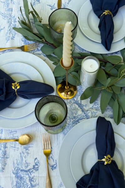 Overhead view of a blue toile tablecloth with navy napkins in gold leaf napkin rings, white plates, green ribbed goblets, gold flatware, and ivory taper candles set into a eucalyptus garland