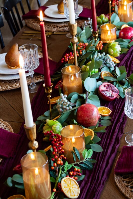 Close-up of a Thanksgiving centerpiece with a halved pomegranate, dried orange slices, red berries, faux artichokes, and amber glass pillar candles nestled in eucalyptus