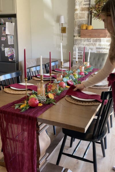 Burgundy gauze napkin being placed on a white plate and woven placemat at a Thanksgiving tablescape with pomegranates, dried orange slices, and burgundy taper candles