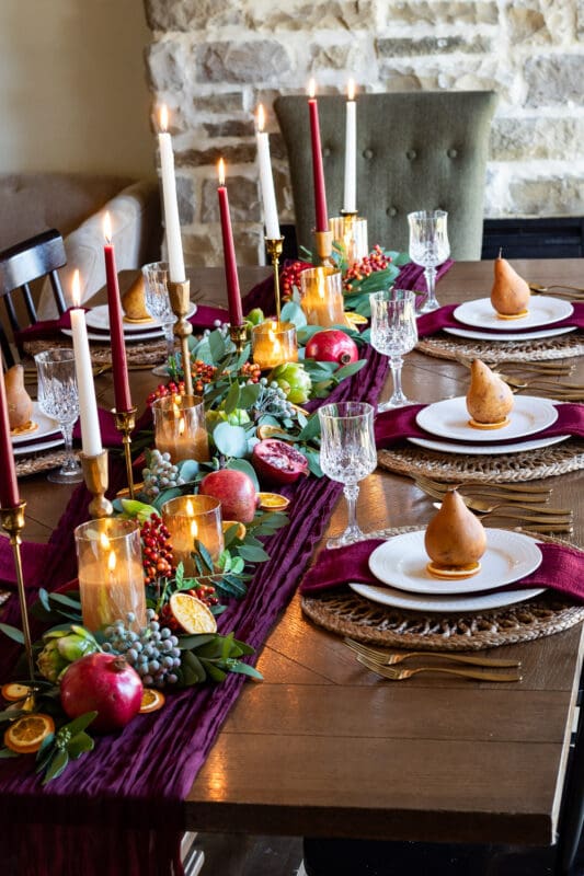 Burgundy Thanksgiving tablescape with a gauze runner, eucalyptus garland, pomegranates, dried orange slices, and burgundy and ivory taper candles, with a golden pear at each place setting