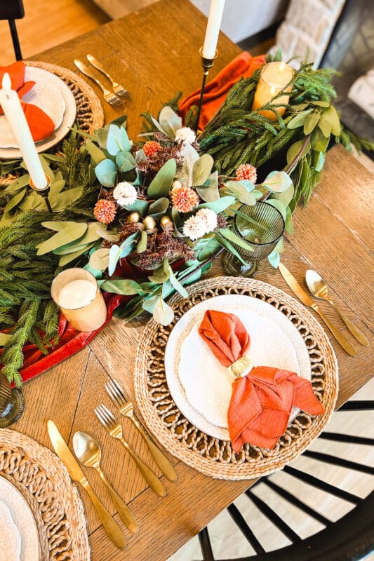 Overhead view of a Thanksgiving place setting with a terracotta napkin in a gold ring on white beaded plates, gold flatware, and a woven placemat beside a centerpiece of eucalyptus, pine, faux mums, and gold acorns