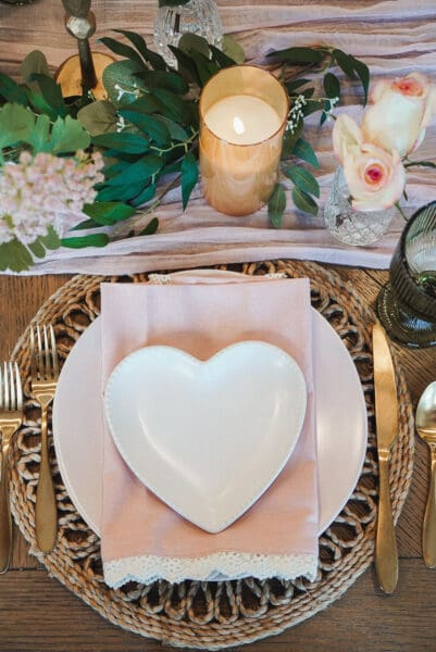 Overhead view of a Valentine's place setting with a white heart-shaped plate on a pink dinner plate, a blush lace-trimmed napkin, gold flatware, and a green ribbed goblet on a round woven placemat