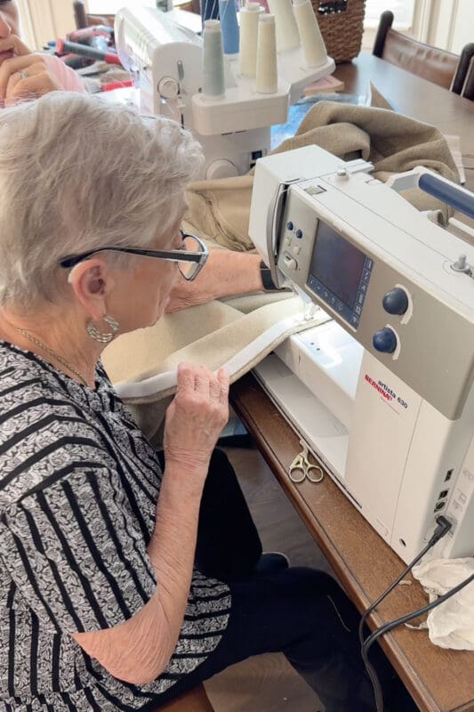 A friend sewing the neutral cushion cover on a Bernina sewing machine at the dining table, with a serger set up behind her