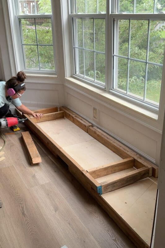 The assembled 2x4 seat frame for the bay window bench with storage resting on the plywood base, with cross braces spaced along its length