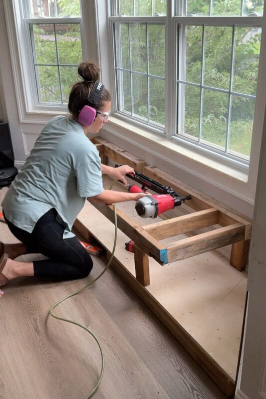 Crystel nailing the raised seat frame into the 2x4 ledger on the wall, building the support structure for the bay window bench with storage