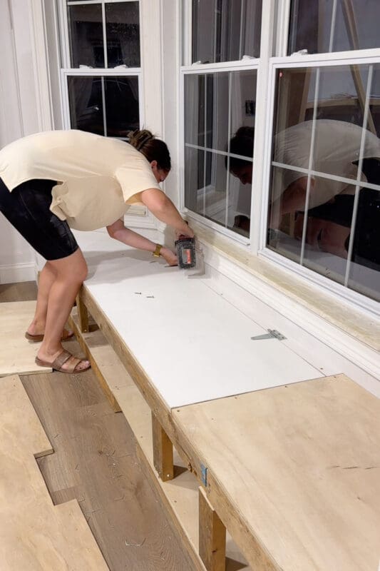 Crystel is using a dril along the back edge of the bench top at night, to attach metal hinges between the fixed back strip and the lift top panel