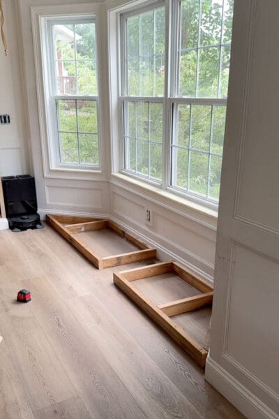 Two wood base frames set in place along the bay window wall with a gap left in the middle for the floor vent, forming the foundation of the storage bench
