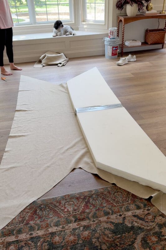 Long strips of upholstery foam taped together next to fabric spread out on the floor to make a cushion for the bay window bench with storage, with a small dog sitting on the bench in the background