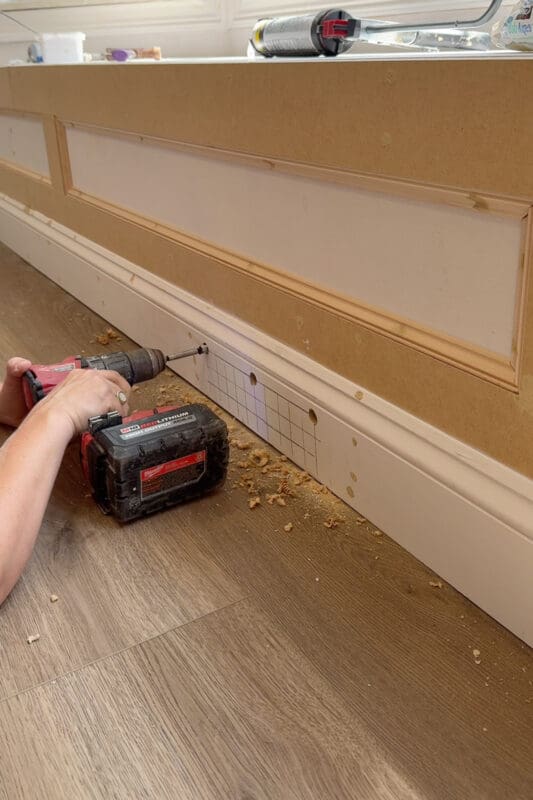 Drilling a grid of round holes into the baseboard at the base of the bench to create a custom air vent, with wood shavings scattered on the floor
