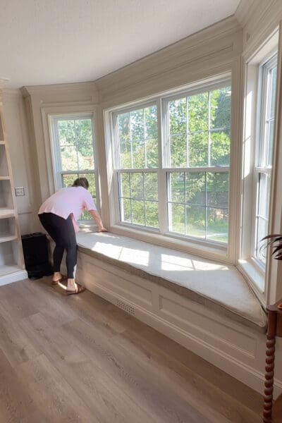 Crystel smoothing the long neutral cushion into place on the finished bay window bench with storage, with the DIY baseboard vent visible below