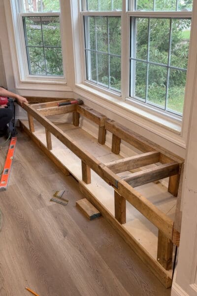 The completed 2x4 framing for the bay window bench with storage, with vertical legs, cross braces, and an open cavity above the plywood base