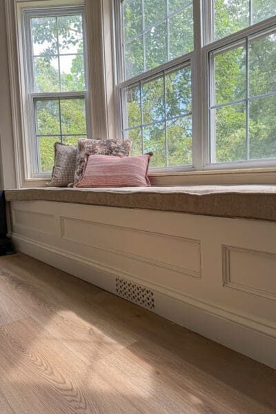 Finished bay window bench with storage painted Accessible Beige, styled with a long neutral cushion, three throw pillows, and the drilled vent visible in the baseboard