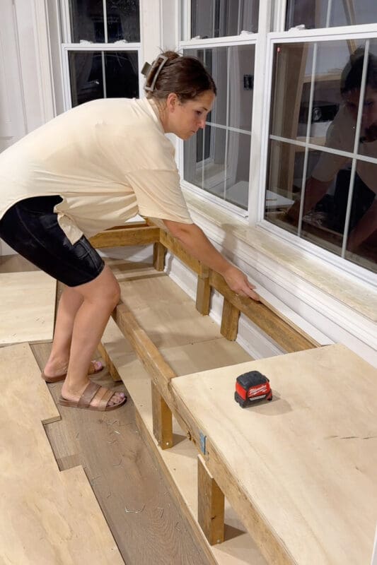 Crystel setting a narrow board along the back of the bench framing at night, with a cut plywood top panel already resting on the right side section