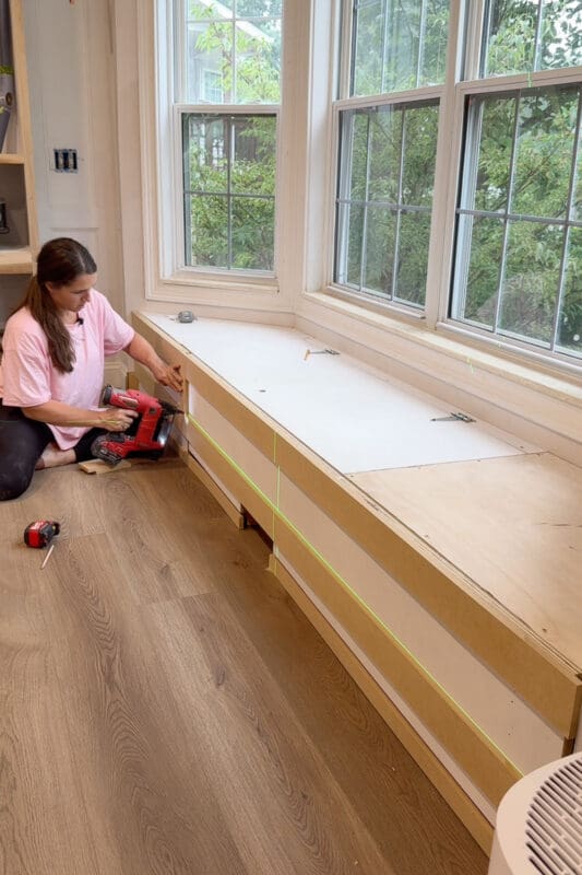 Crystel kneeling with a brad nailer to attach trim pieces to the front of the bay window bench with storage, with a green laser line running along the panel