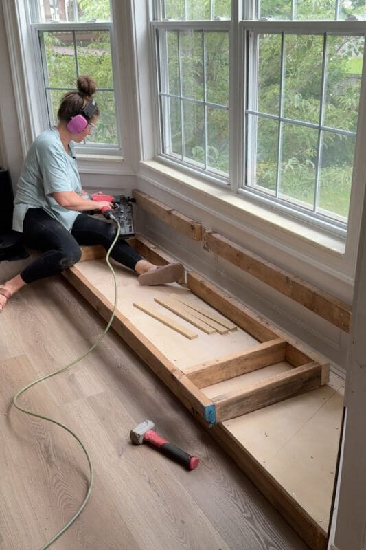 Crystel kneeling with a tape measure to check the seat frame position, with a green laser level line running across the wall behind her