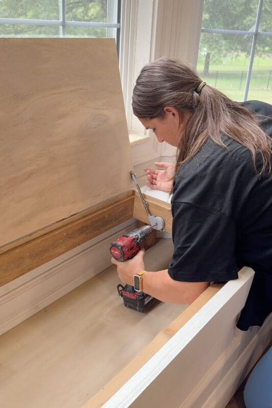 Crystel holding the bench lid open with one hand while positioning a metal lid support hinge inside the storage cavity