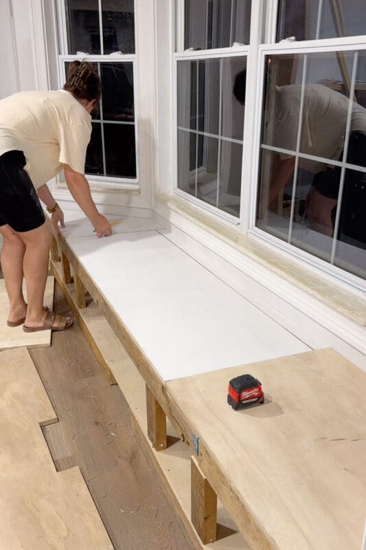 Crystel marking a cut line with a pencil and straightedge on the white faced panel that will become the opening lift top for the bay window bench with storage