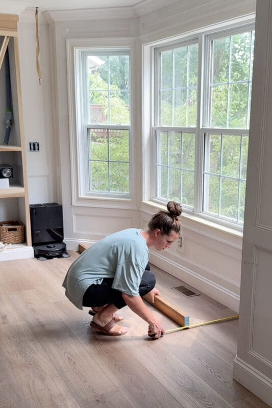 Crystel crouching on the floor with a tape measure and a wood board, measuring the bay window nook to plan the bay window bench with storage