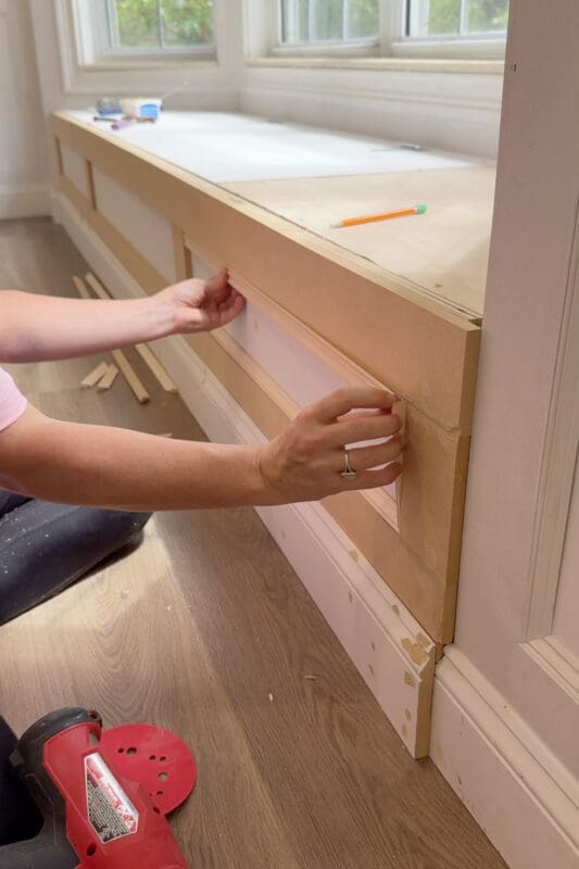 Two hands holding a thin trim strip in place on the bench front to build a picture frame molding pattern that matches the existing wall trim