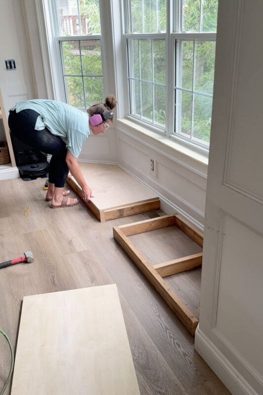 Crystel setting a sheet of plywood over the wood base frame in the bay window nook, with the floor vent visible in the open middle section