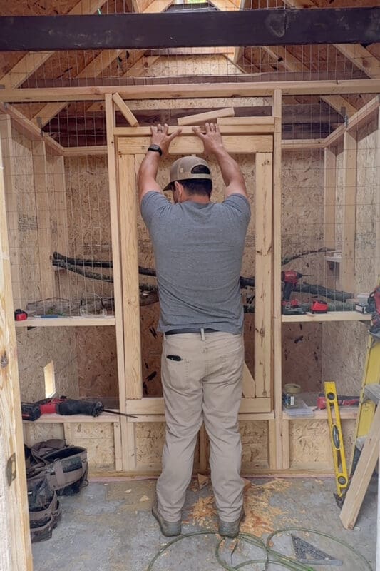 Man holding a framed wire gate up in the opening of the center dividing wall that separates the chicken roosting area from the storage side