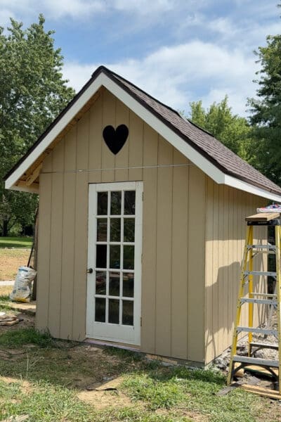 Finished tan chicken coop with brown shingled roof, white trim, heart shaped gable cutout, and a vintage glass paned door