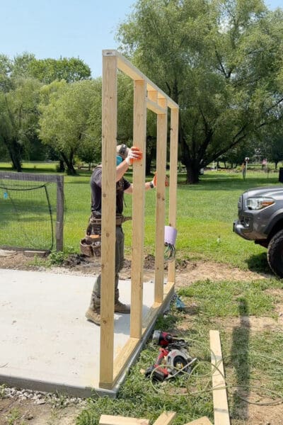 Man standing up the first framed 2x4 wall on the concrete slab while building an outdoor home for backyard hens, with a circular saw and drill on the grass nearby
