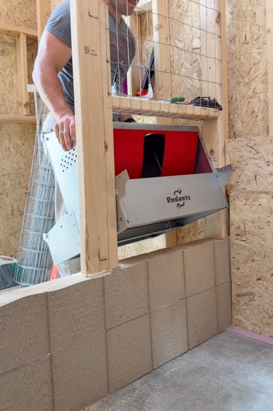 Man fitting a red and metal roll away nesting box into a framed opening in the chicken coop dividing wall from the roosting side