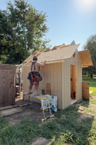 Man on a work platform nailing a 2x4 sub fascia board across the cut rafter tails of a chicken coop while OSB roof sheathing goes on above