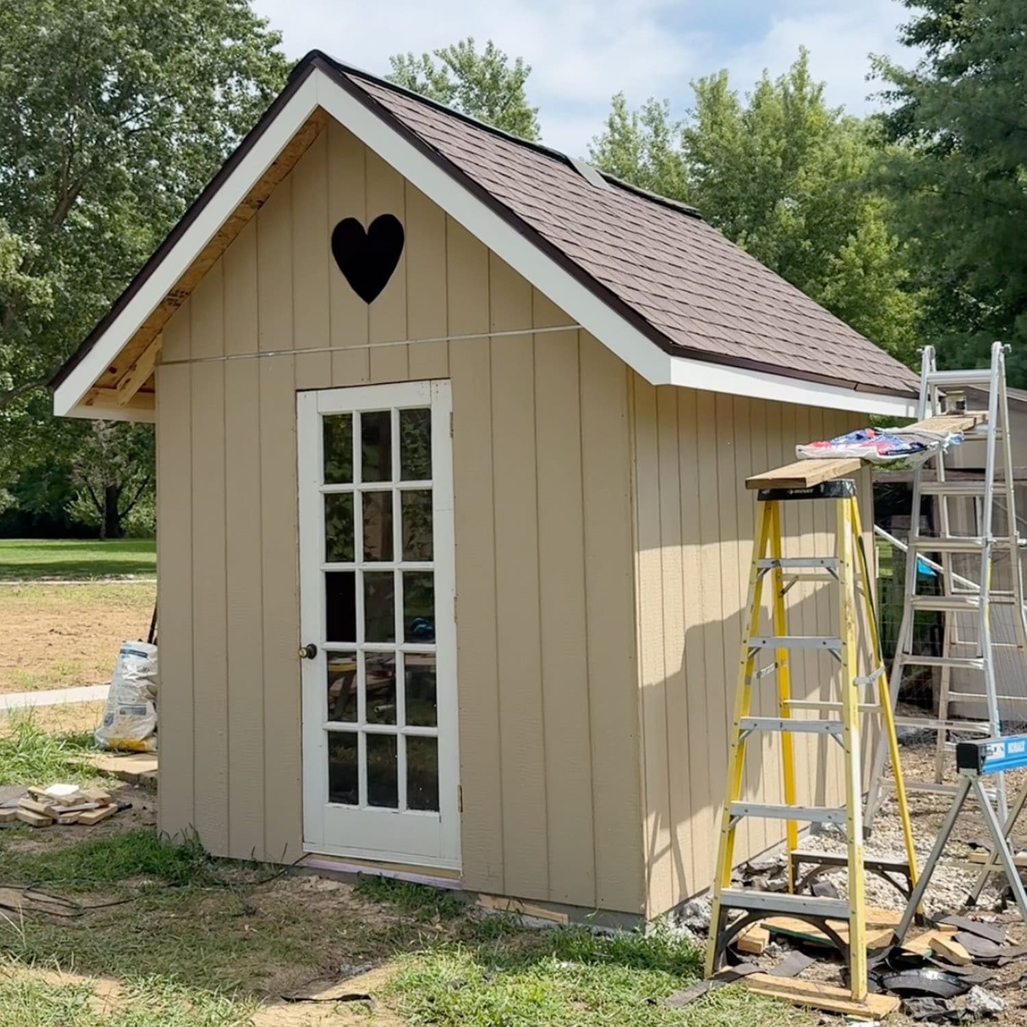 Woman in a pink striped tee posing with arms out in front of a tan DIY chicken coop with a heart cutout in the gable and a vintage glass-paned door