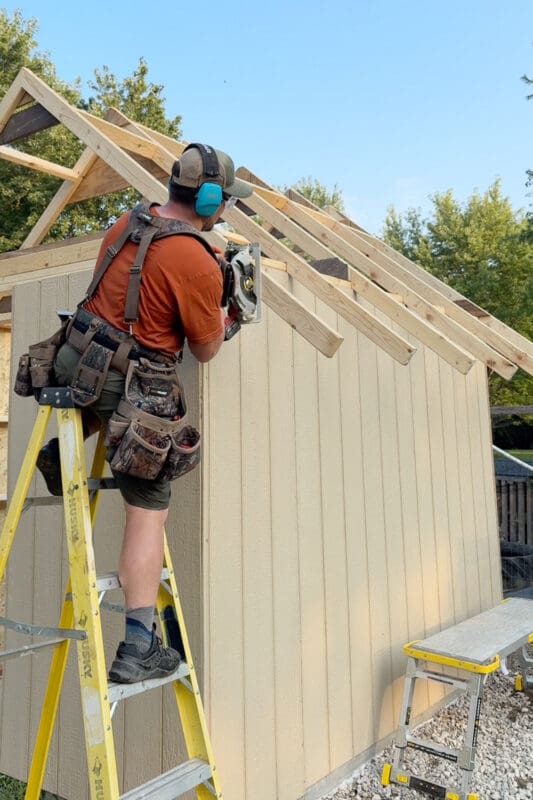 Man on a ladder using a circular saw to trim the top edge of chicken coop siding along the angled roof line