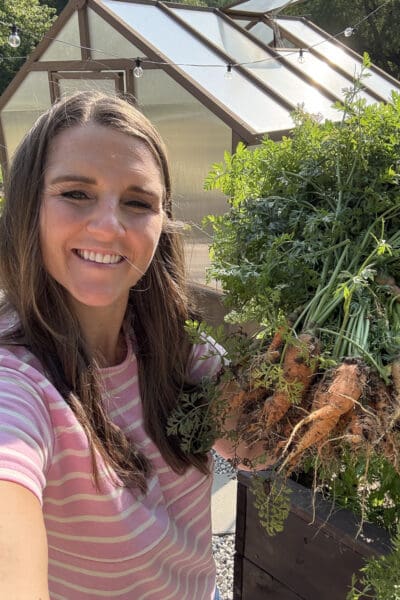 Crystel holding a bunch of freshly pulled garden carrots with the leafy greens still attached, standing in front of her greenhouse