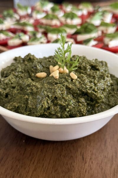 Close-up of carrot top pesto in a white bowl garnished with pine nuts and a carrot green frond, served alongside a caprese platter