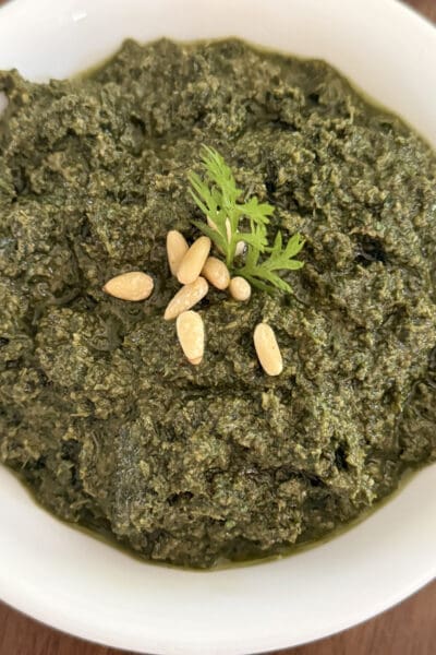 Overhead view of carrot top pesto showing its thick, coarse texture and deep green color, finished with pine nuts and a carrot leaf sprig