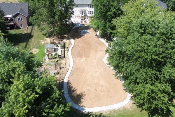 Aerial view of the finished concrete path in the backyard curving in a full loop around a large bare dirt area, with the house, patio, and greenhouse visible at the edges