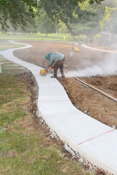 Worker using a walk-behind concrete saw to cut relief joints across the cured concrete path in the backyard, with dust rising and red marking lines showing where each cut goes