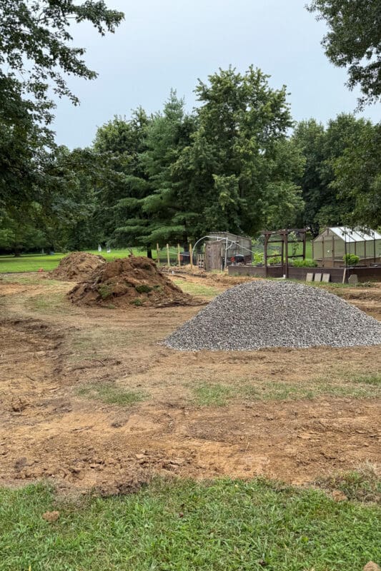 Pile of excavated dirt next to a pile of gravel road base in the leveled backyard, with the greenhouse and garden beds in the background